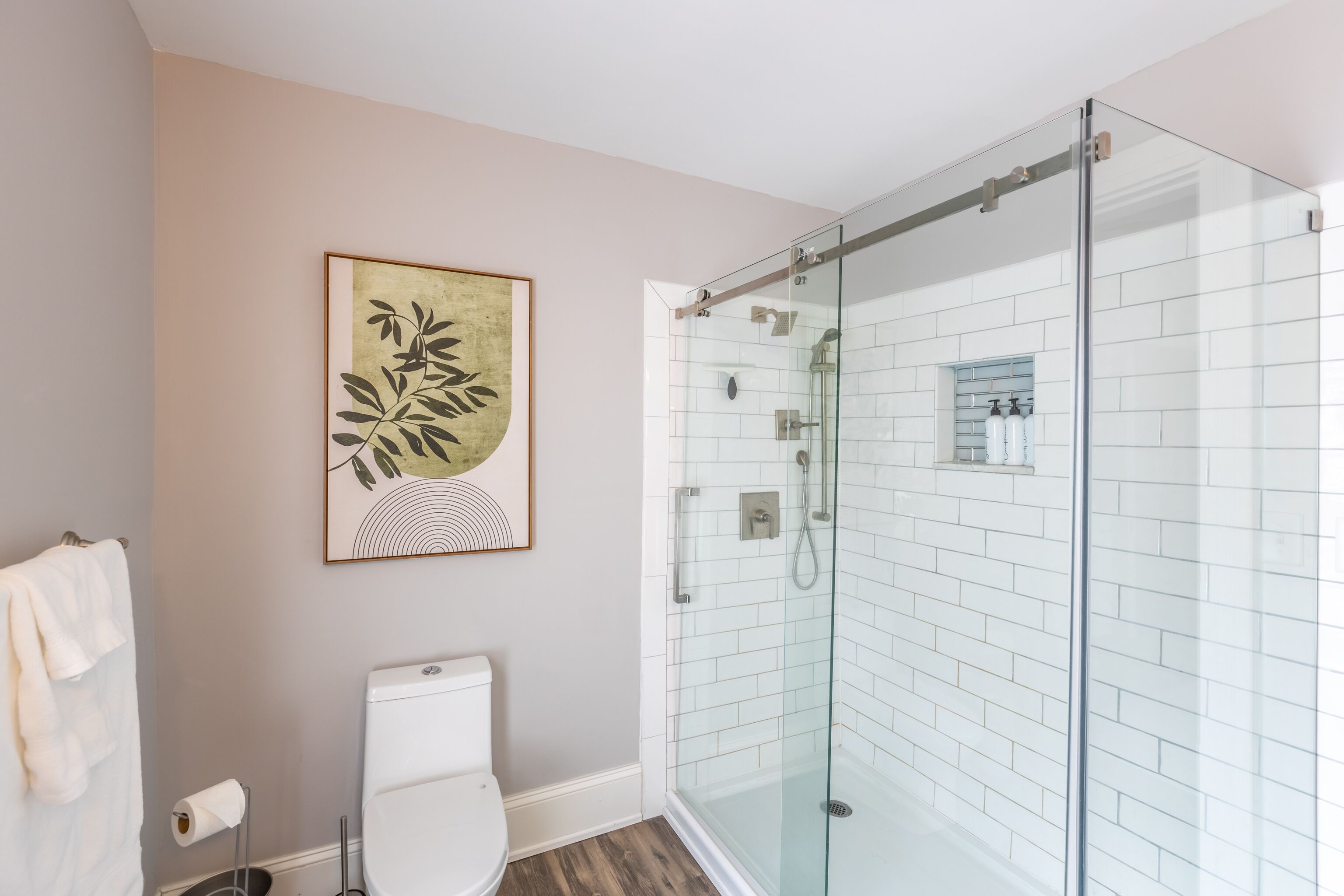 Un baño con ducha separada por vidrio, inodoro y una obra de arte enmarcada en la pared.