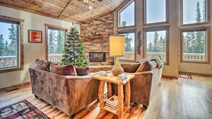 A cozy living room with a fireplace, a Christmas tree, and large windows overlooking a forest. - 22STR-00367 Fairplay Cabin ~ 6 Mi to Downtown! (Fairplay)