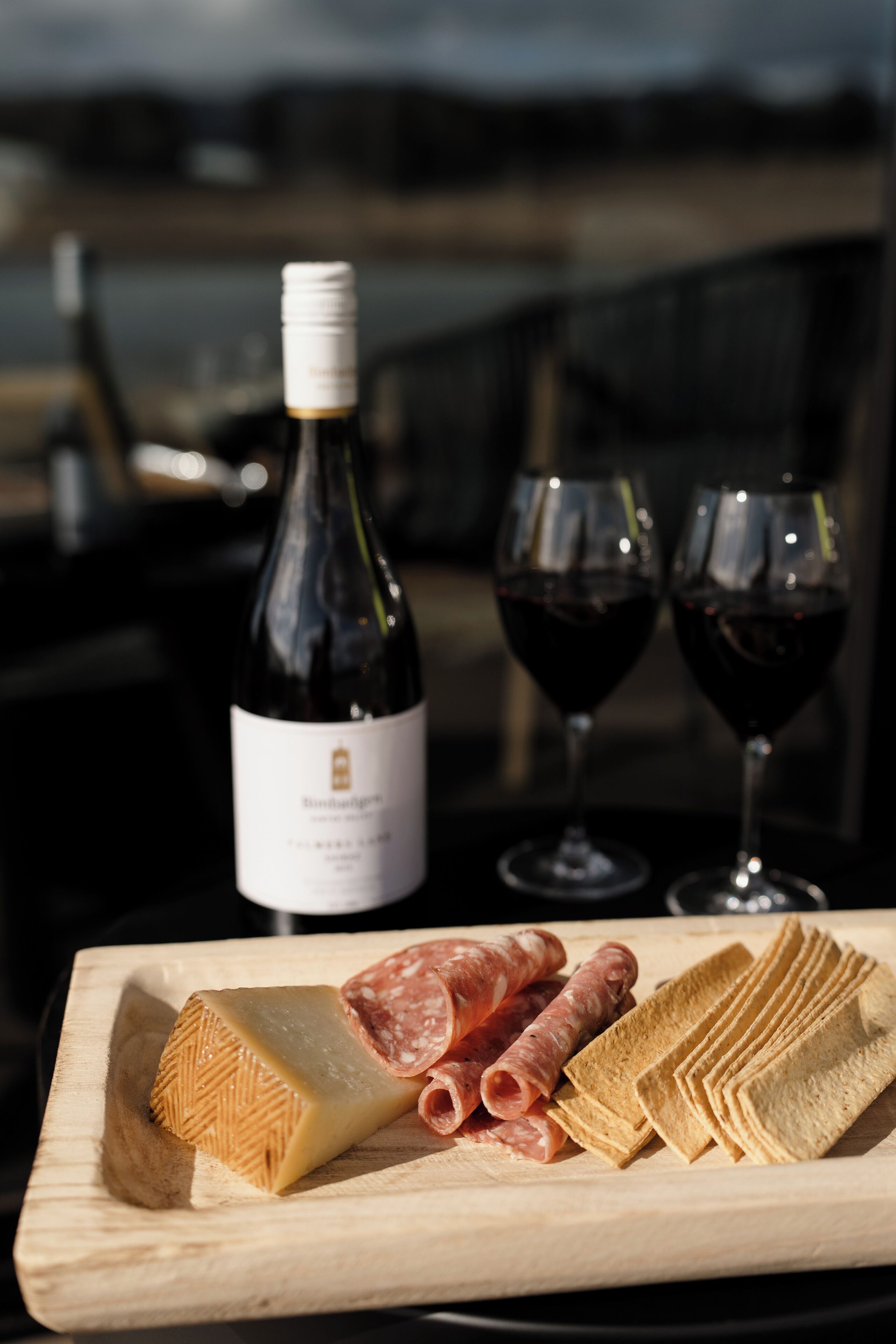 Una taula de tall de fusta amb una llesca de formatge, làmines de salami i galetes. Al fons, hi ha una ampolla de vi i dues copes de vi vermell.
