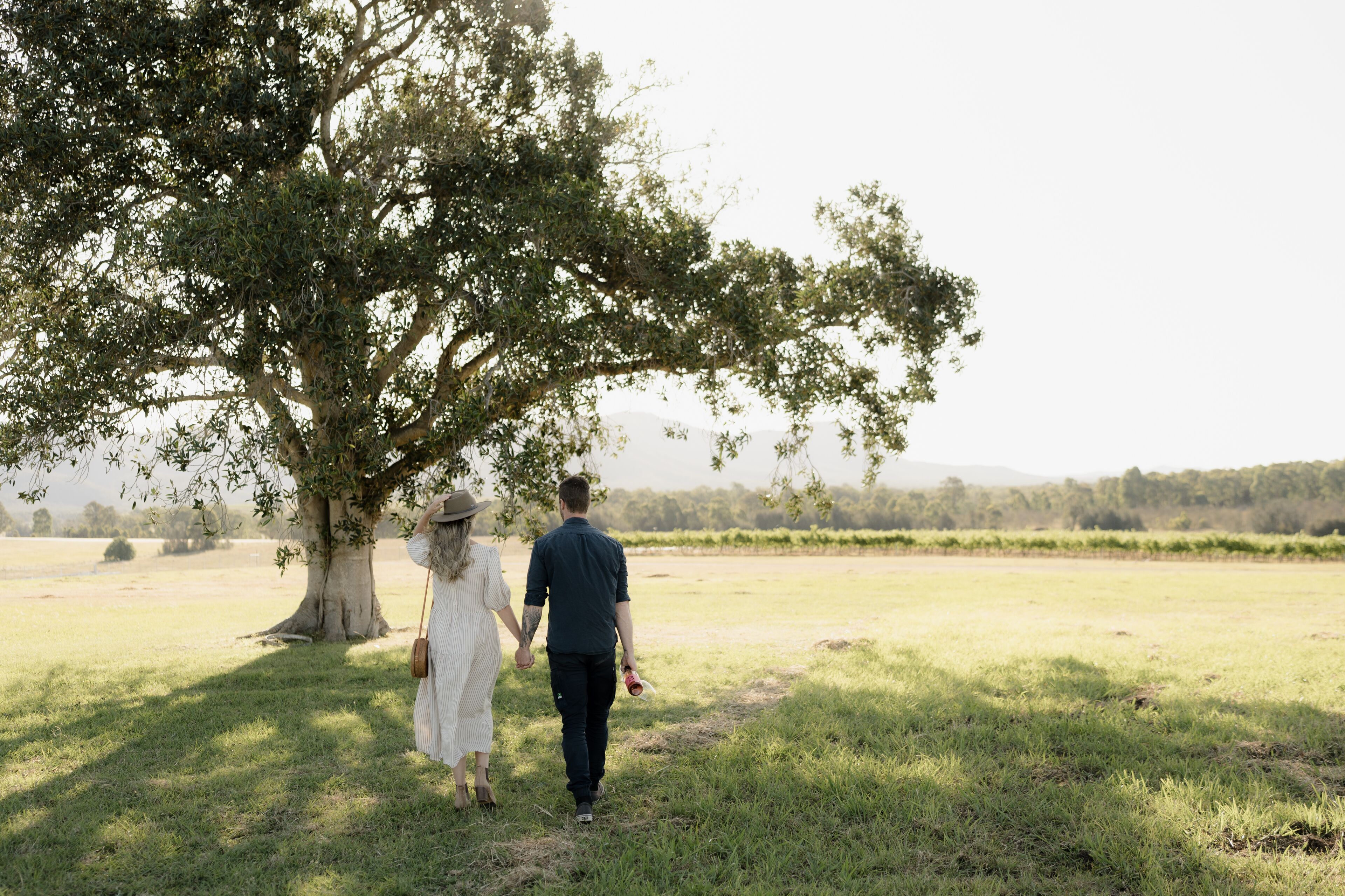 A couple walking under a large tree in a grassy field.