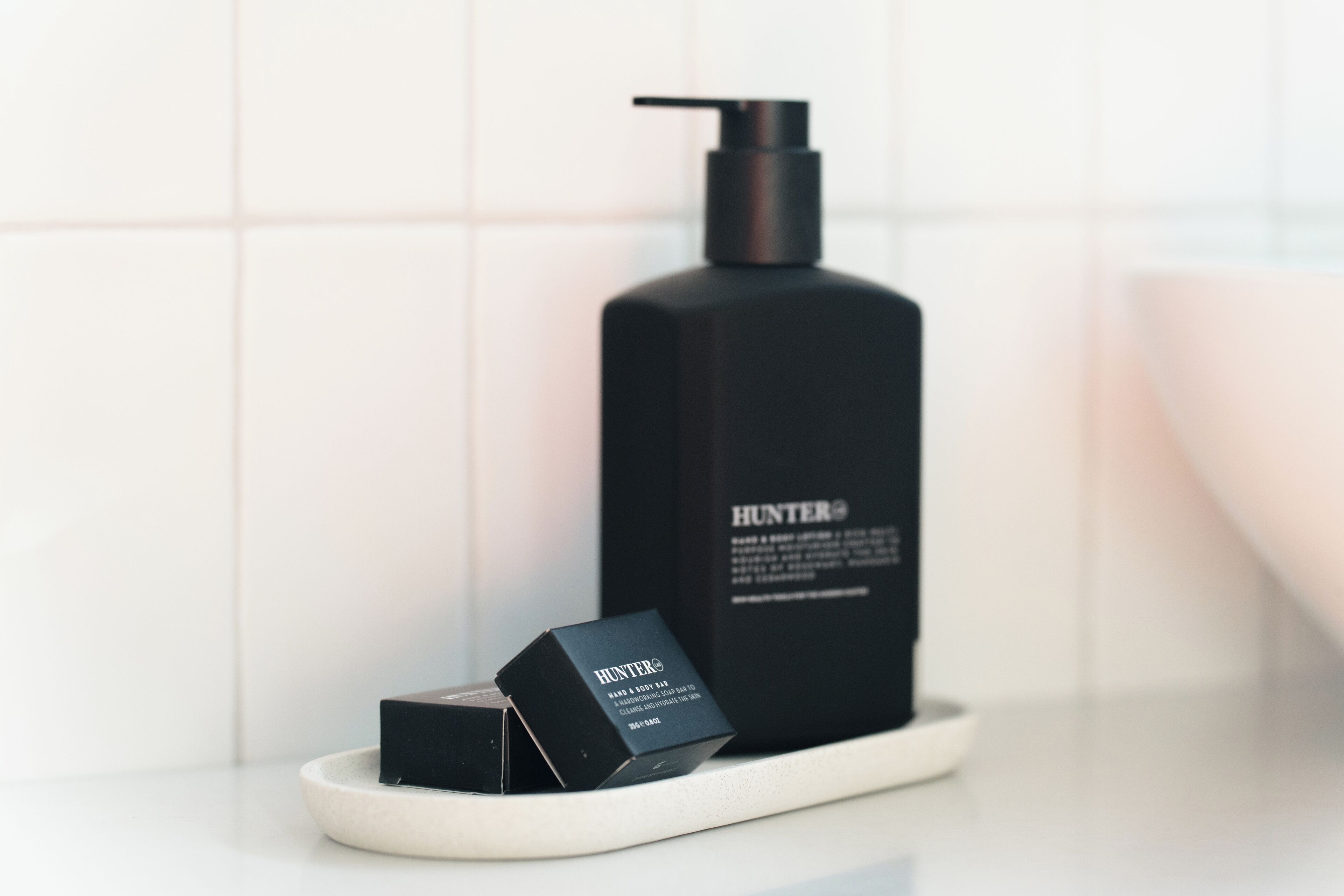 Een zwarte fles Hunter® body lotion op een witte schaal in een badkamer met witte tegels.