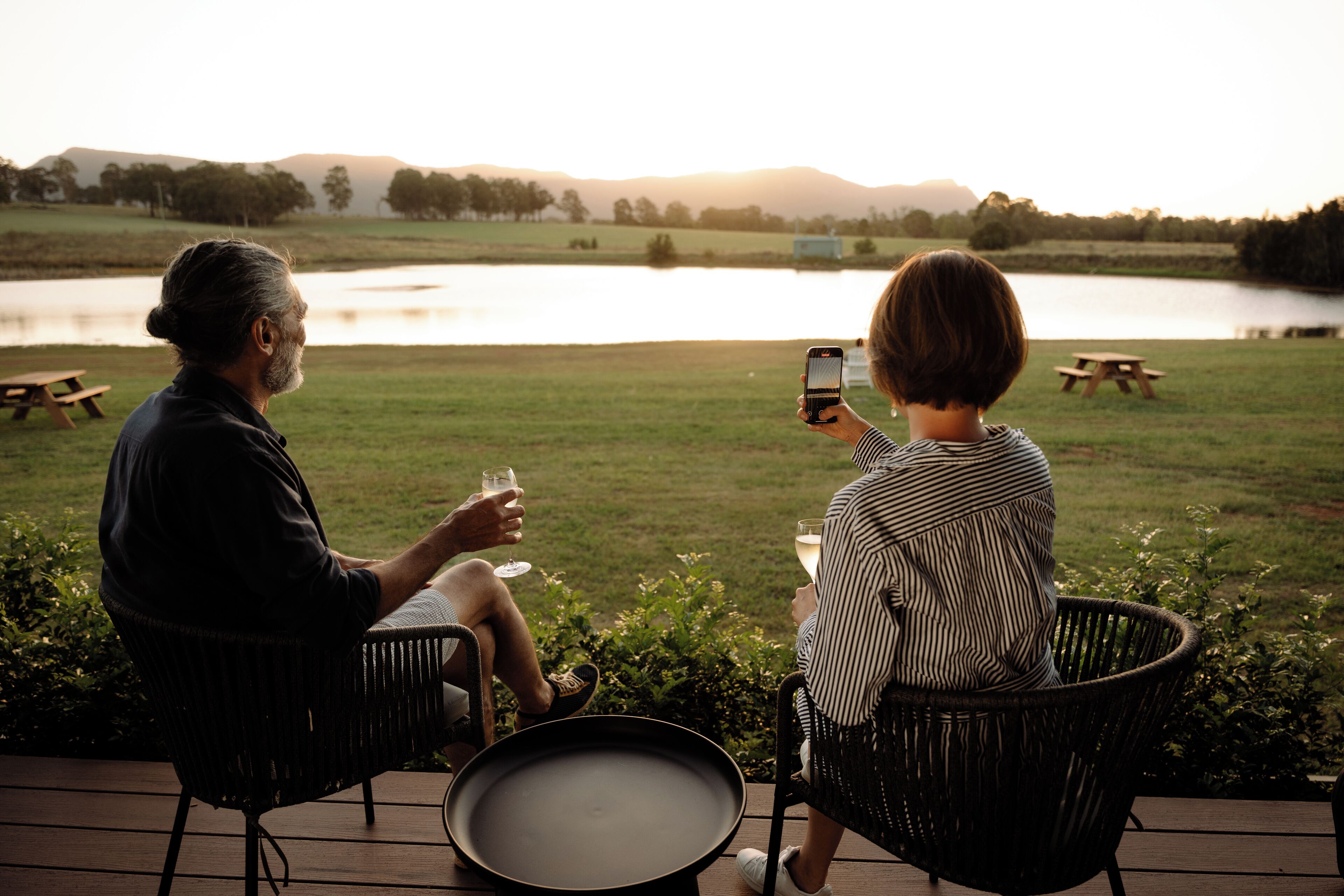 Two people sitting on wicker chairs facing a body of water with picnic tables in the background.