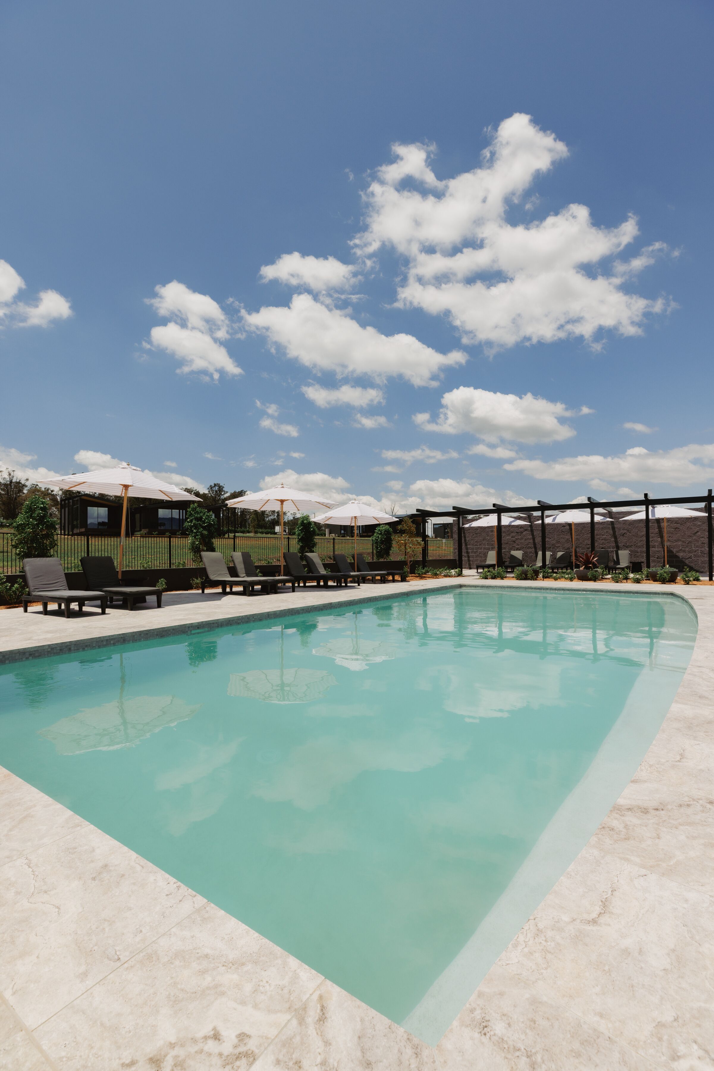 A swimming pool with lounge chairs and umbrellas, surrounded by a fence and greenery.