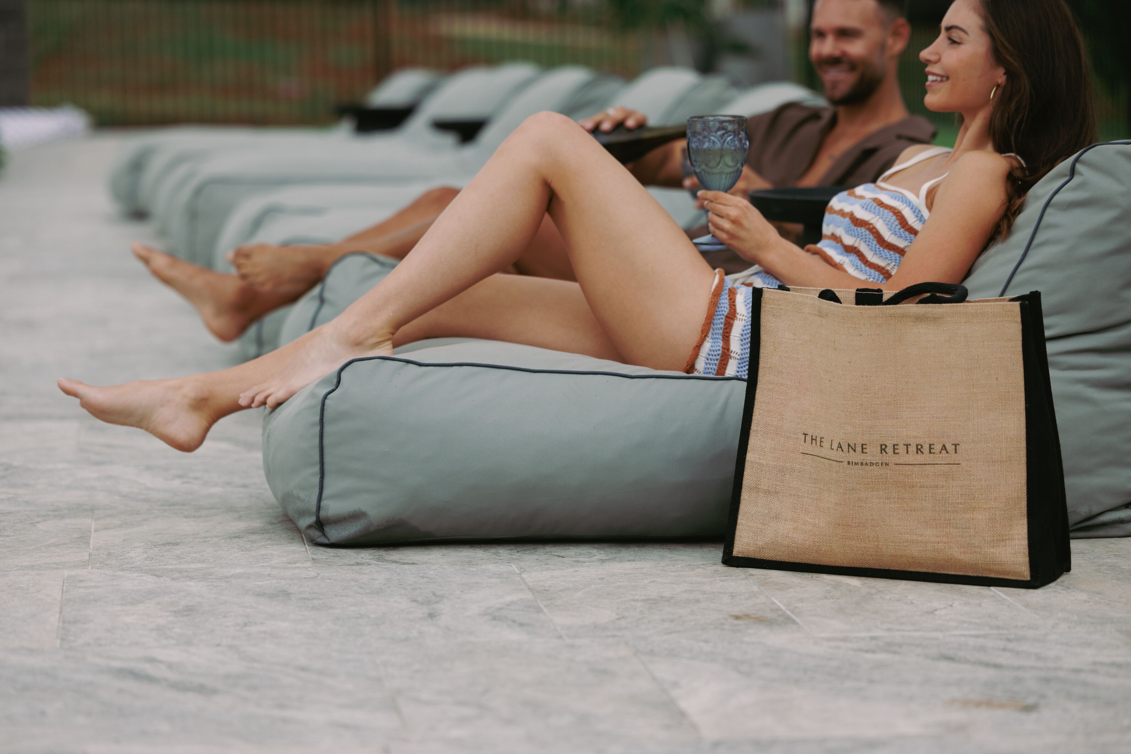 Una pareja descansando en sillones de exterior con vista a la playa, sosteniendo bebidas y una bolsa con la etiqueta 'The Lane Retreat'.