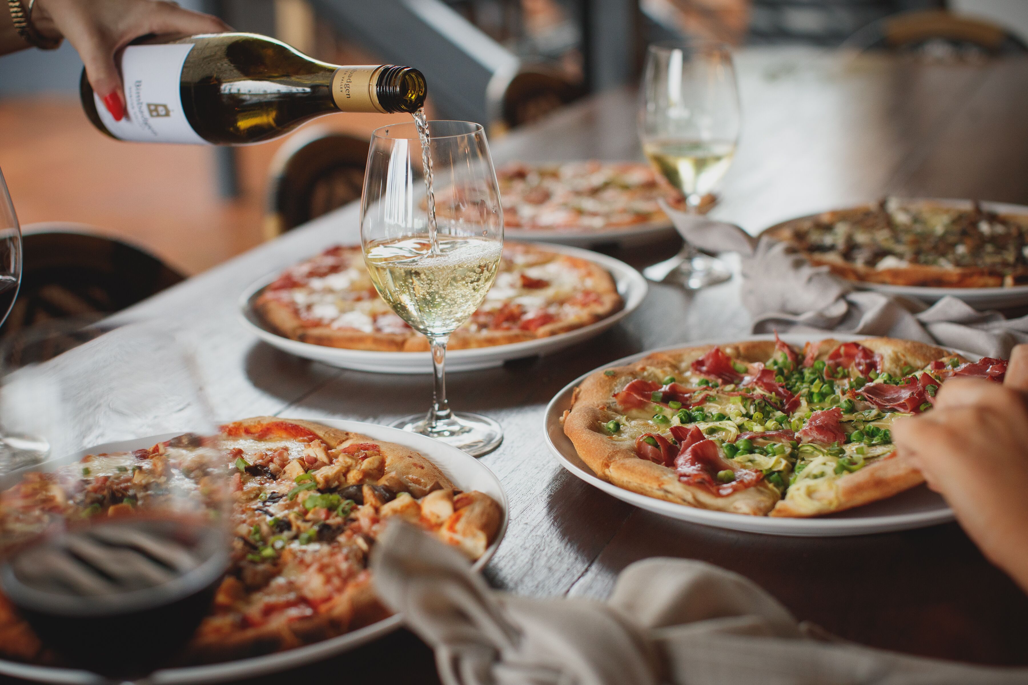Een eettafel met meerdere pizza's en glazen witte wijn.