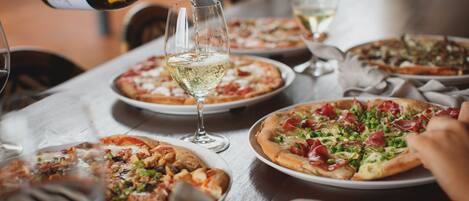 Een eettafel met meerdere pizza's en glazen witte wijn.