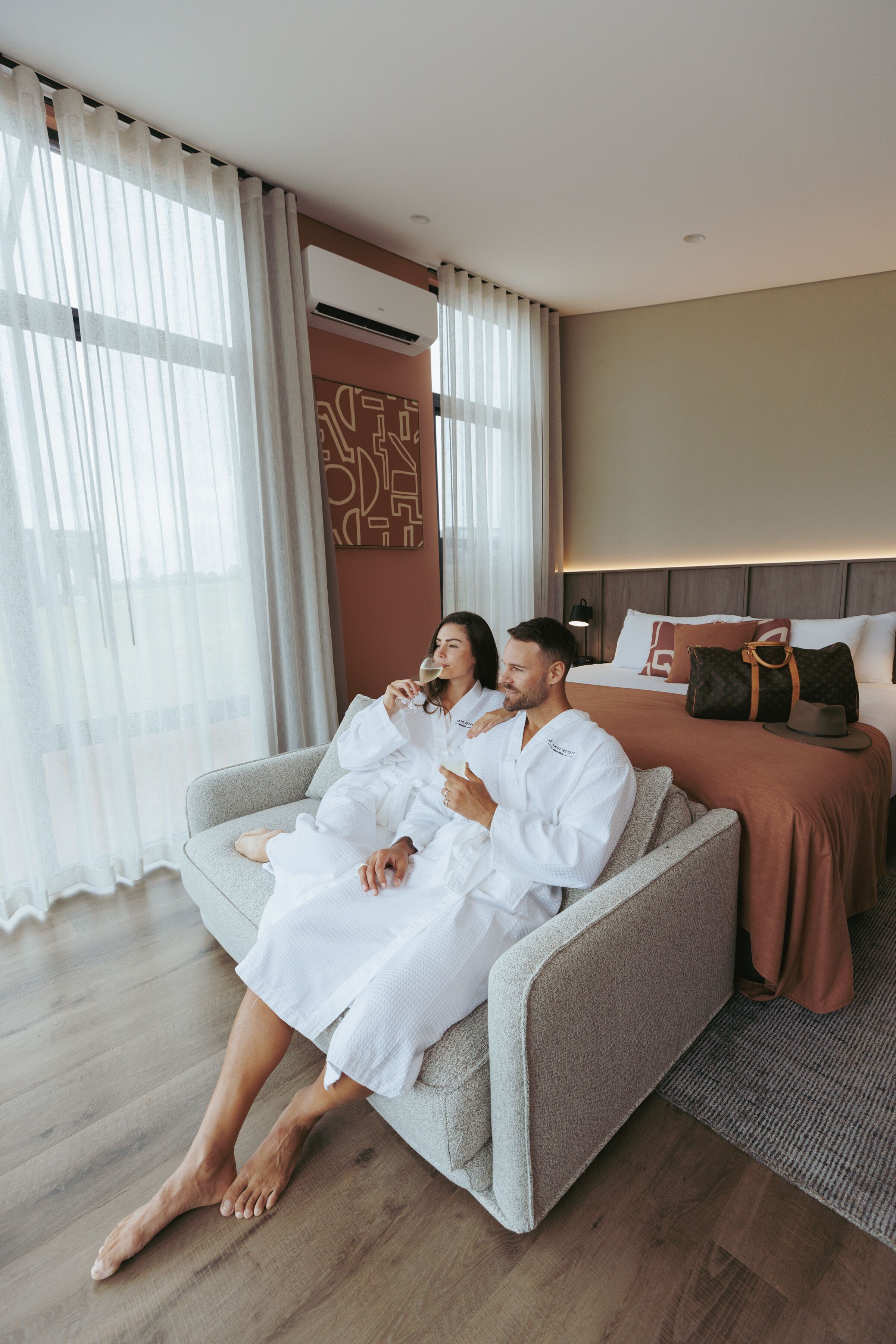 A couple in white robes sitting on a grey sofa in a modern hotel room with a bed in the background.