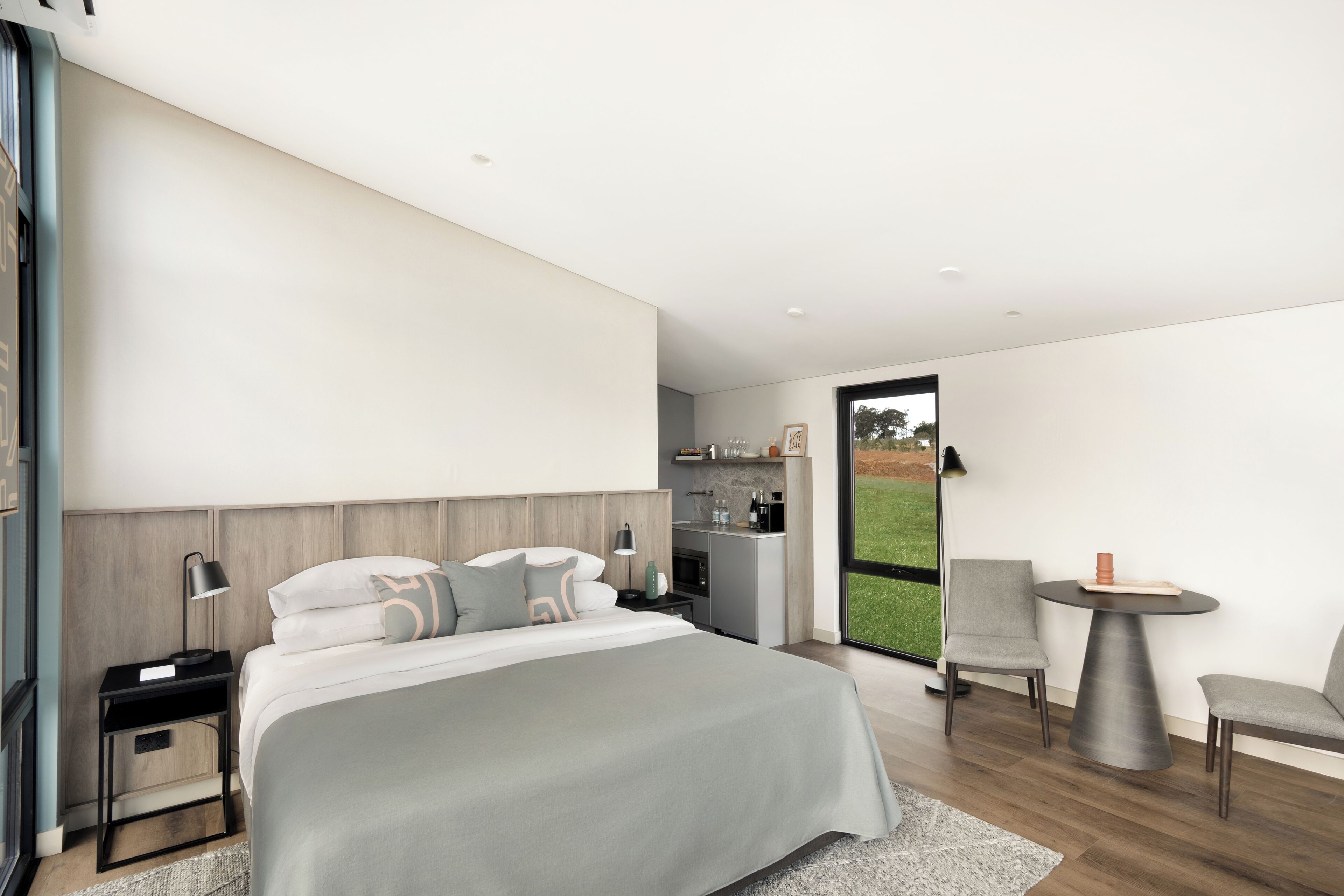 A modern bedroom with a large bed, bedside tables, a small kitchenette, and a seating area with a table and chairs.