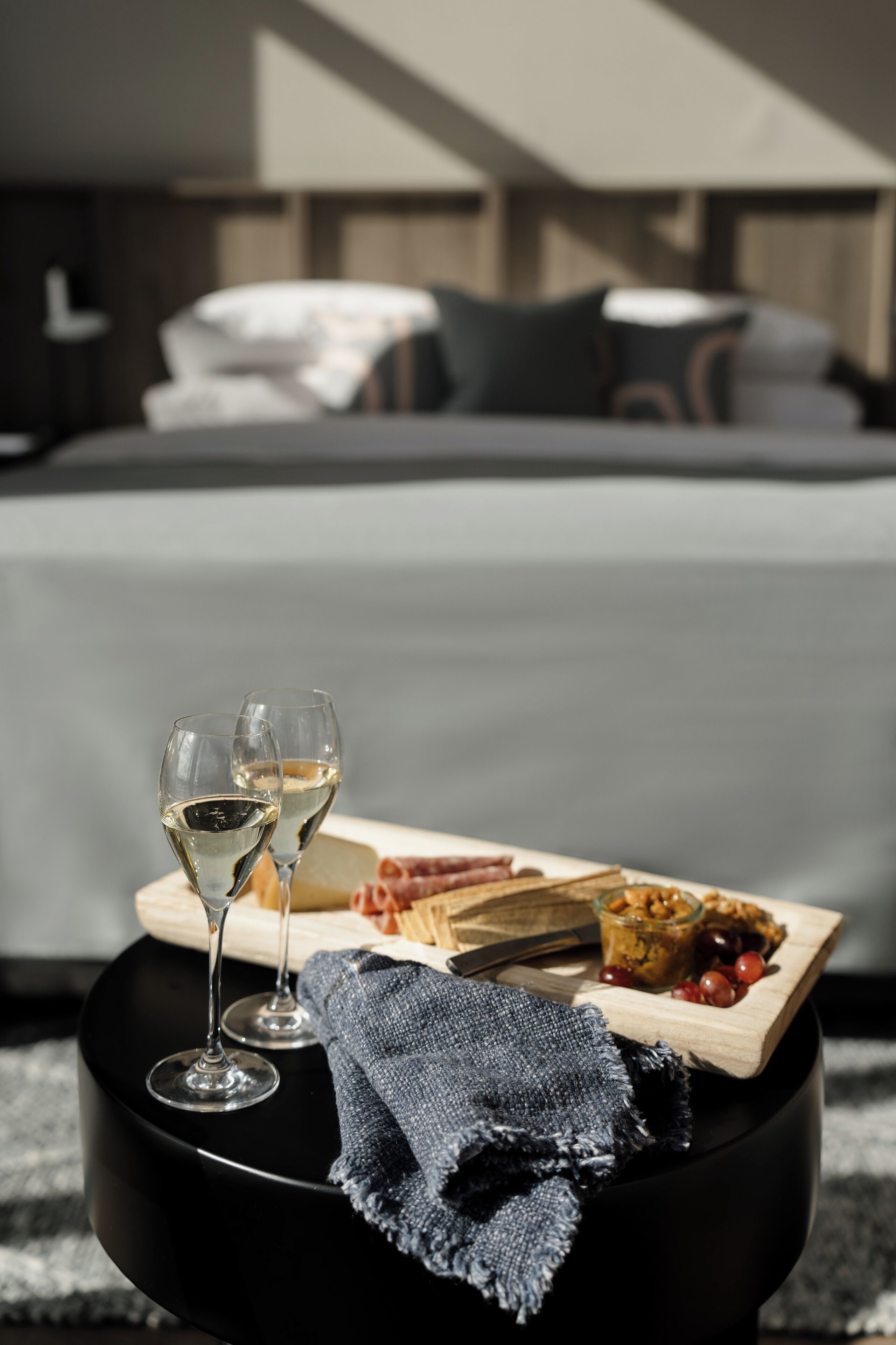 Ein ordentlich angerichteter Frühstückswagen mit zwei Weingläsern, einer Käseplatte und einer kleinen Schale Obst auf einem schwarzen Ottoman in einem modernen Hotelzimmer, im Hintergrund ein Bett.