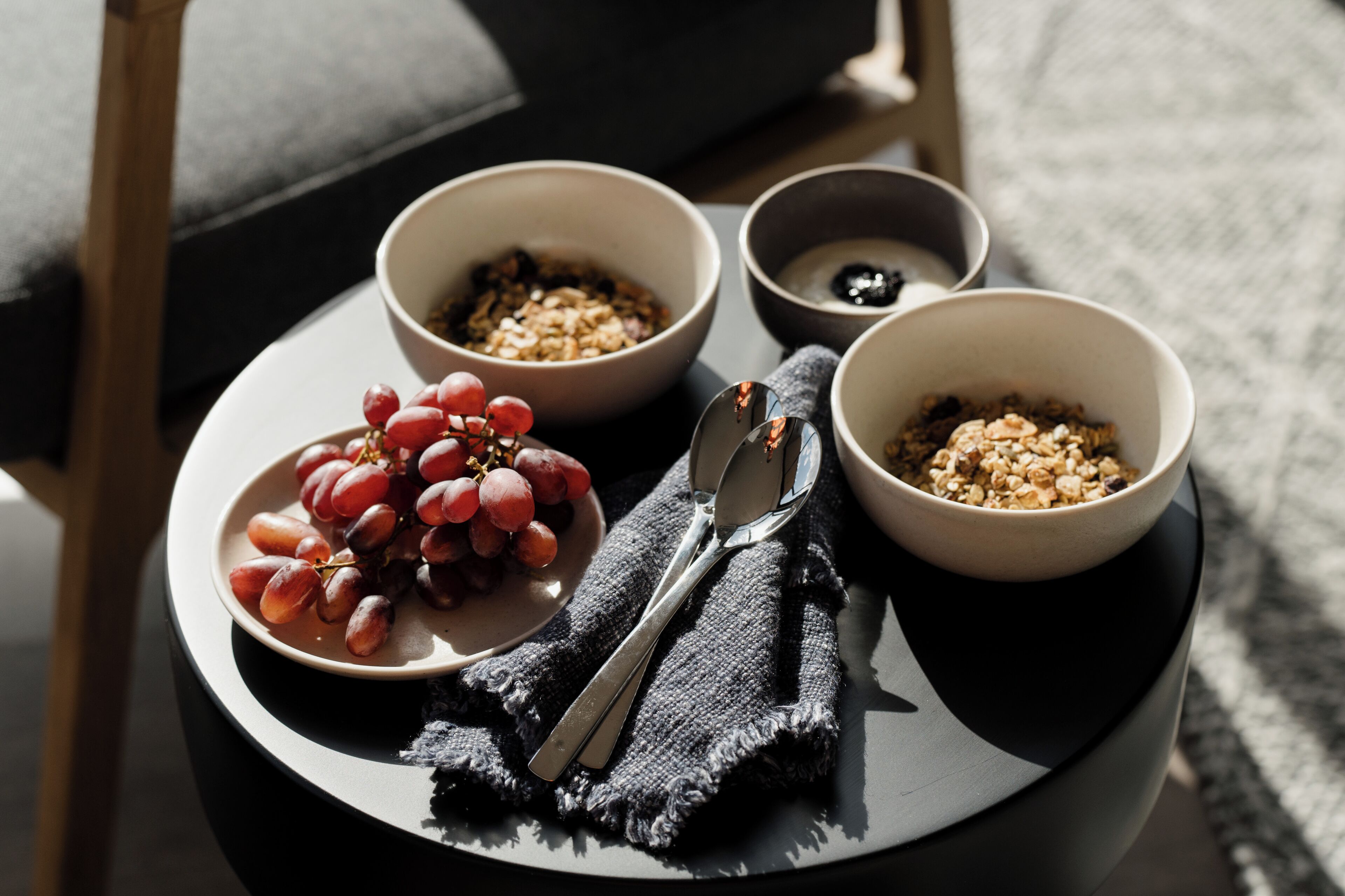 A breakfast tray with bowls of cereal, grapes, and a small bowl of yogurt, placed on a round table with a dark cloth and silverware.