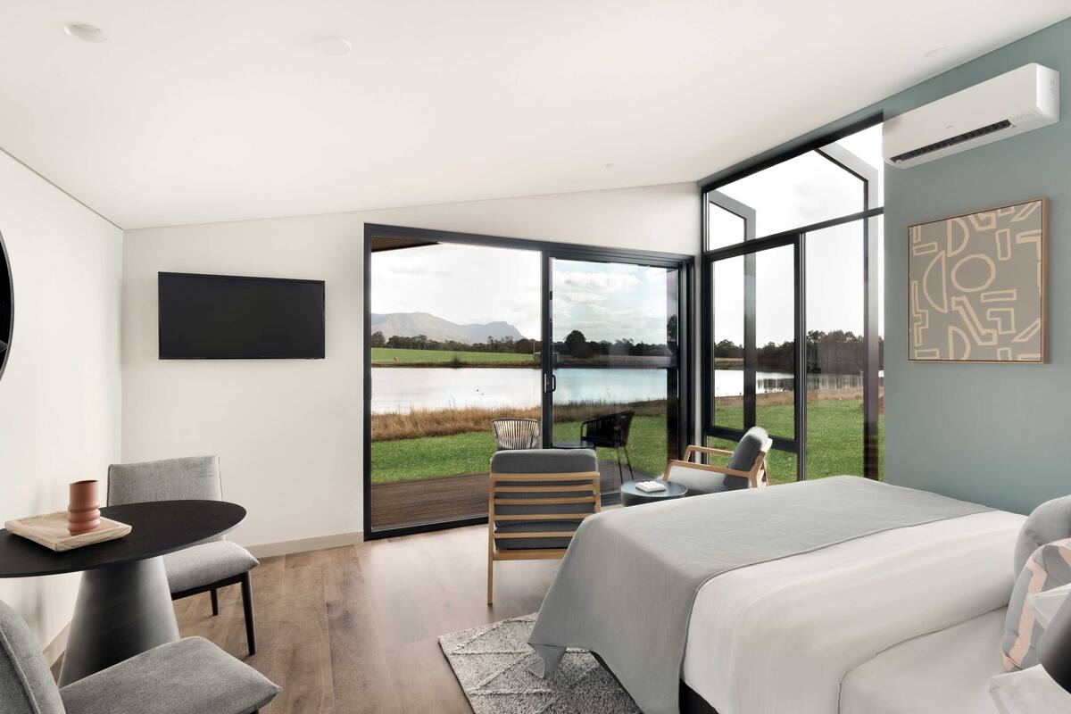 Un dormitorio moderno con una cama grande, un escritorio y una silla. Cuenta con un ventanal con vistas a un lago y montañas.