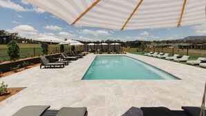 Piscina con sillones de descanso y sombrillas, rodeada de un área ajardinada con árboles y vistas a colinas distantes.