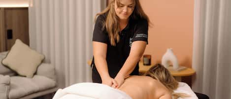 A massage therapy session in a room with a bed, a small table with a lamp, and a couch in the background.