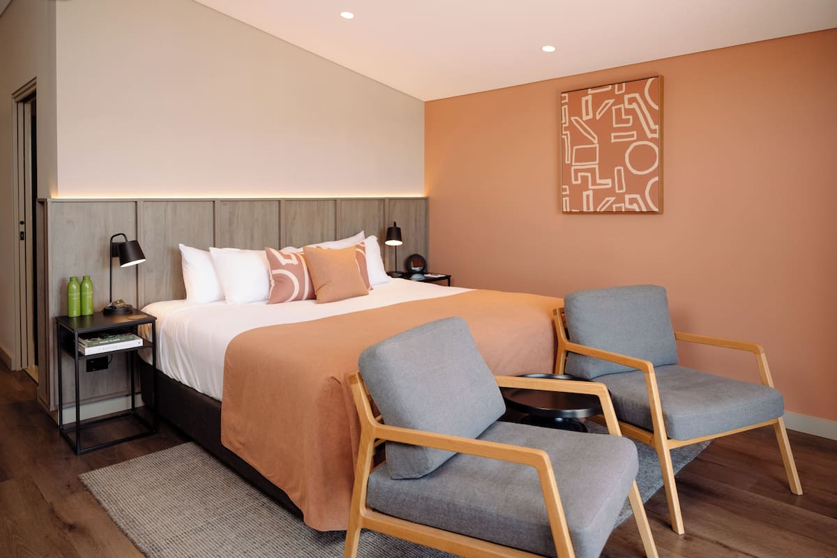 Una habitación de hotel moderna con una cama grande, dos sillones, una mesita de noche con lámpara y una obra de arte en la pared.