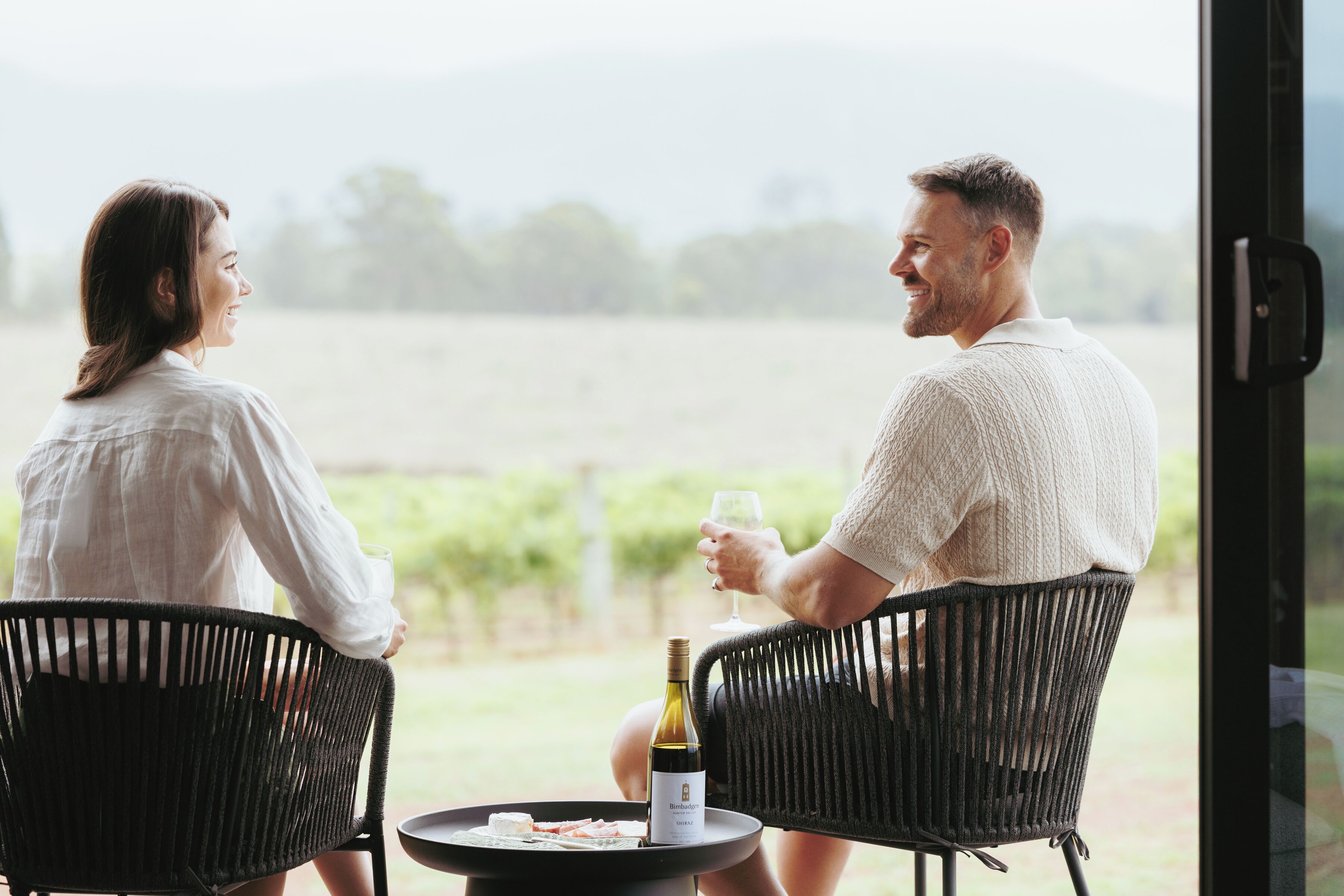 Two people sitting at a table with a bottle of wine and glasses, outdoors with a view of a field.