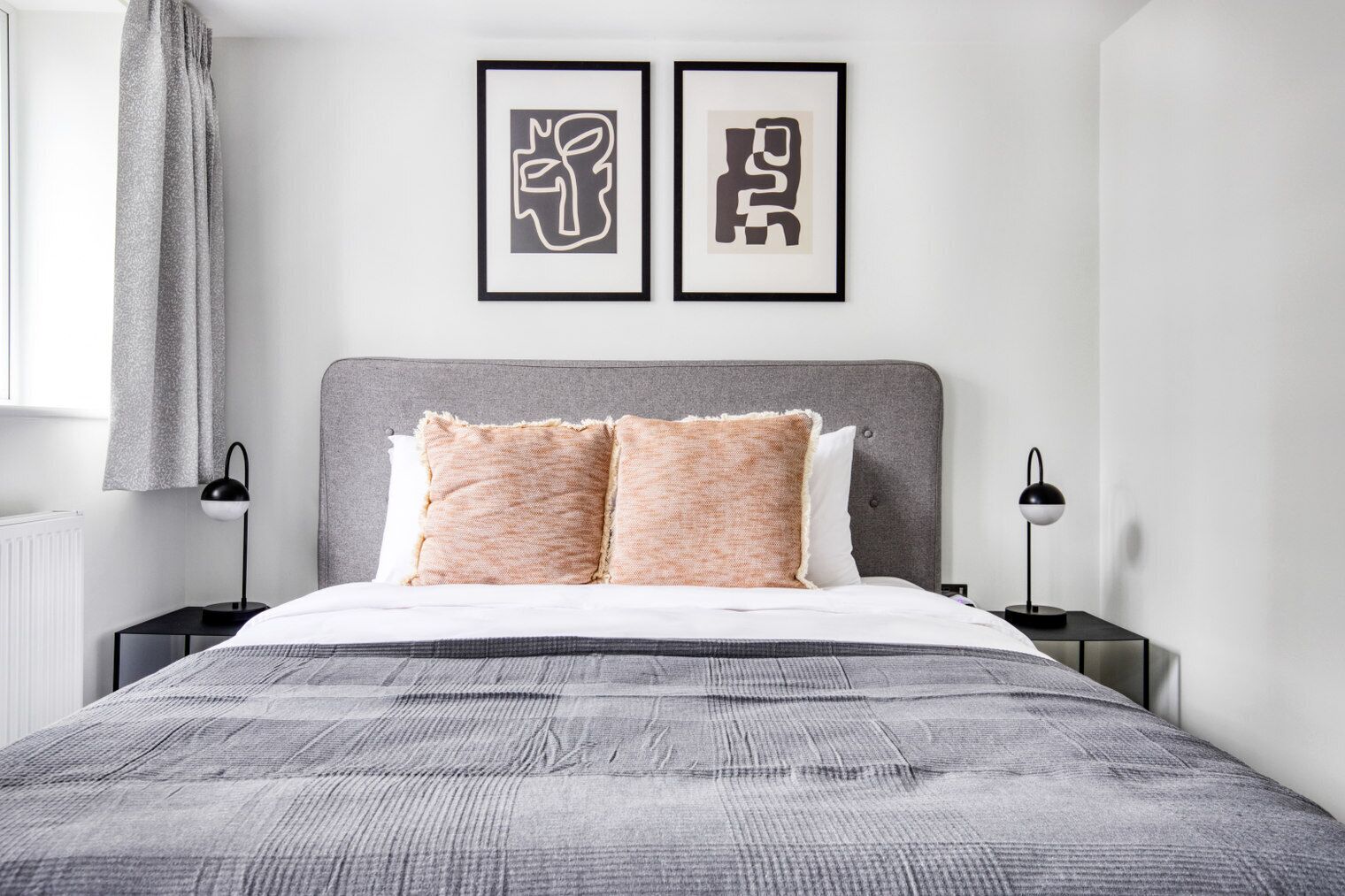 Une chambre moderne avec une tête de lit grise, deux œuvres d’art abstraites encadrées, un couvre-lit gris texturé et deux lampes de chevet.