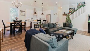 A modern living room with grey sofas, a dining area with a wooden table and chairs, and a staircase with a Christmas tree. - Old Town Bluffton Beaches and Birdies House (Bluffton)