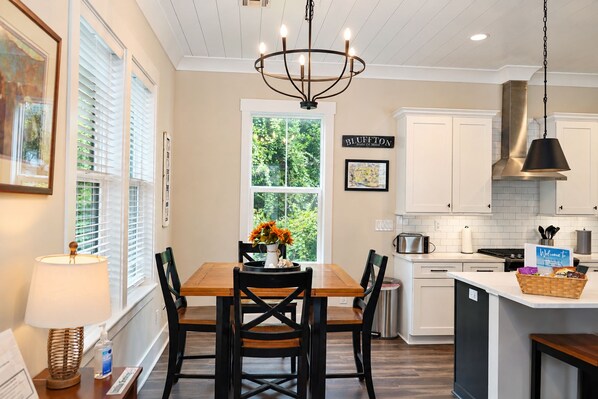 A kitchen with white cabinets, a wooden dining table with chairs, a chandelier, and a window with blinds.