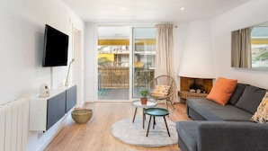 A modern living room with a flat-screen TV, a sofa, a chair, a small table, and a sliding glass door leading to an outdoor area.