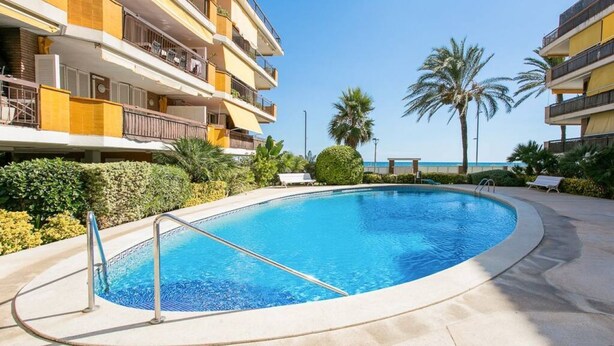 A swimming pool surrounded by a building with balconies, palm trees, and a clear sky.