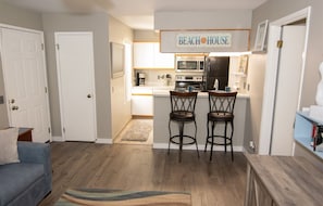 Cozinha compacta com micro-ondas, forno e máquina de café, dois banquetas altas e um letreiro com a inscrição 'BEACH HOUSE'.
