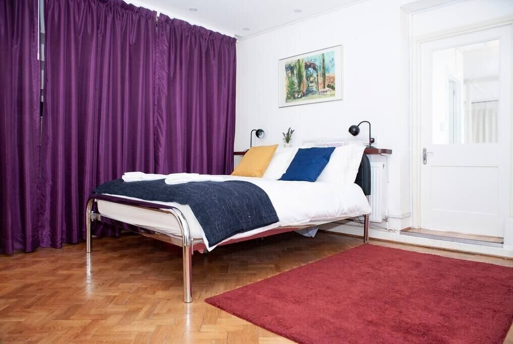 A neatly made bed with a red rug, a purple curtain, and a painting on the wall.