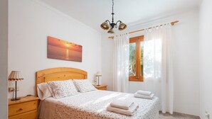 A neatly made bed with a floral bedspread, two bedside lamps, a wooden nightstand, a painting of a sunset, and a window with white curtains.
