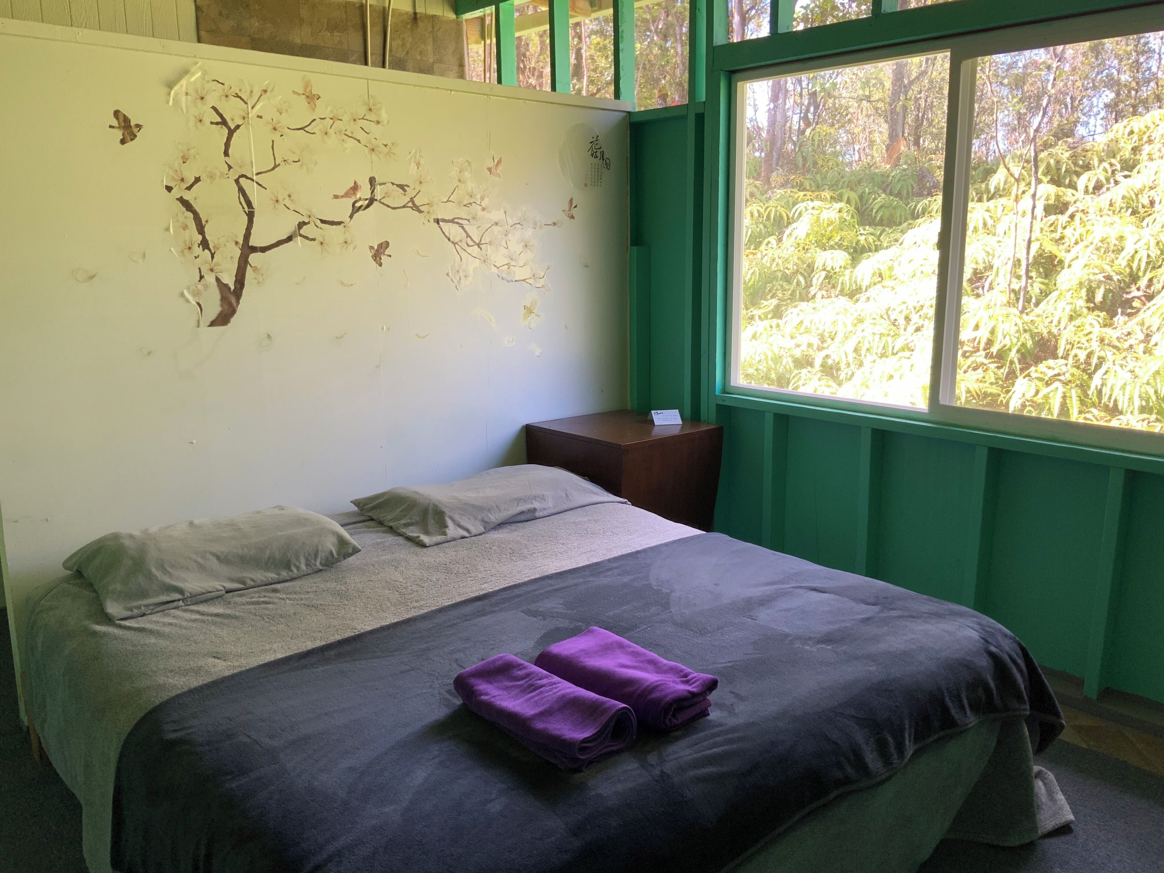 Une chambre avec un lit, une fenêtre donnant sur un paysage verdoyant et un mur décoré d’un arbre et de papillons.