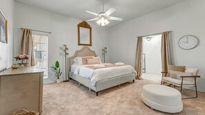 Un dormitorio con cama, una silla, ventilador de techo, reloj y ventana con cortinas.