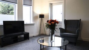 A modern living room with a flat-screen TV on a black stand, a glass coffee table, a grey armchair, and a vase with orange flowers.