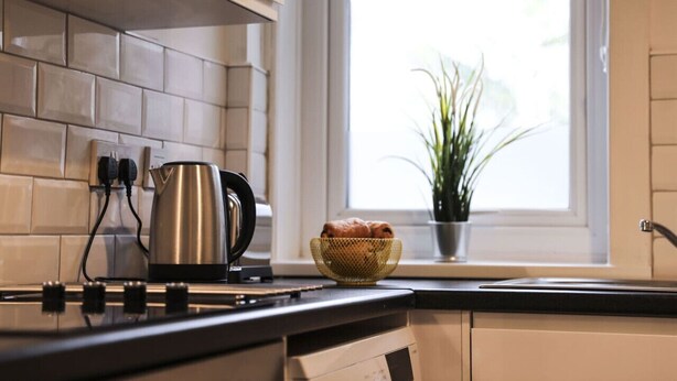 A kitchen with a tiled backsplash, a kettle, a basket of bread, and a window with a plant.