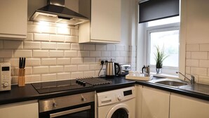 A modern kitchen with white cabinetry, a stainless steel range hood, a built-in oven, a washing machine, and a window with a view.