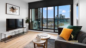 A modern living room with a large TV, a comfortable sofa, a small round table, and a large window offering a city view. - Lumina Suites (West Melbourne)