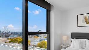 A hotel room with a large window offering a city view, a neatly made bed, a bedside table with a lamp, and a framed artwork on the wall. - Lumina Suites (West Melbourne)