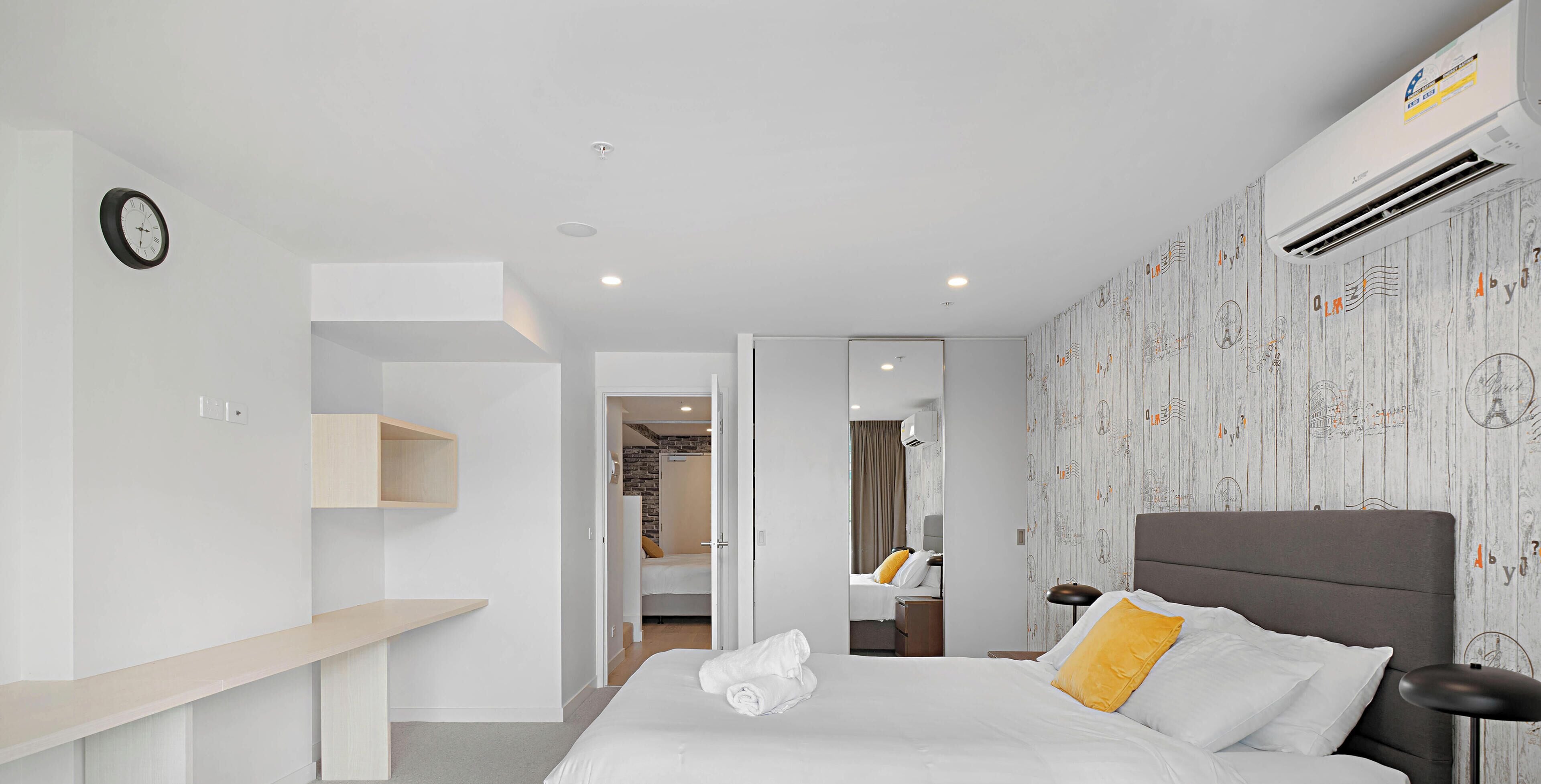 Une chambre d’hôtel moderne, dotée d’un grand lit, d’une table de chevet, d’une armoire et d’une salle de bain visible à travers une porte ouverte.
