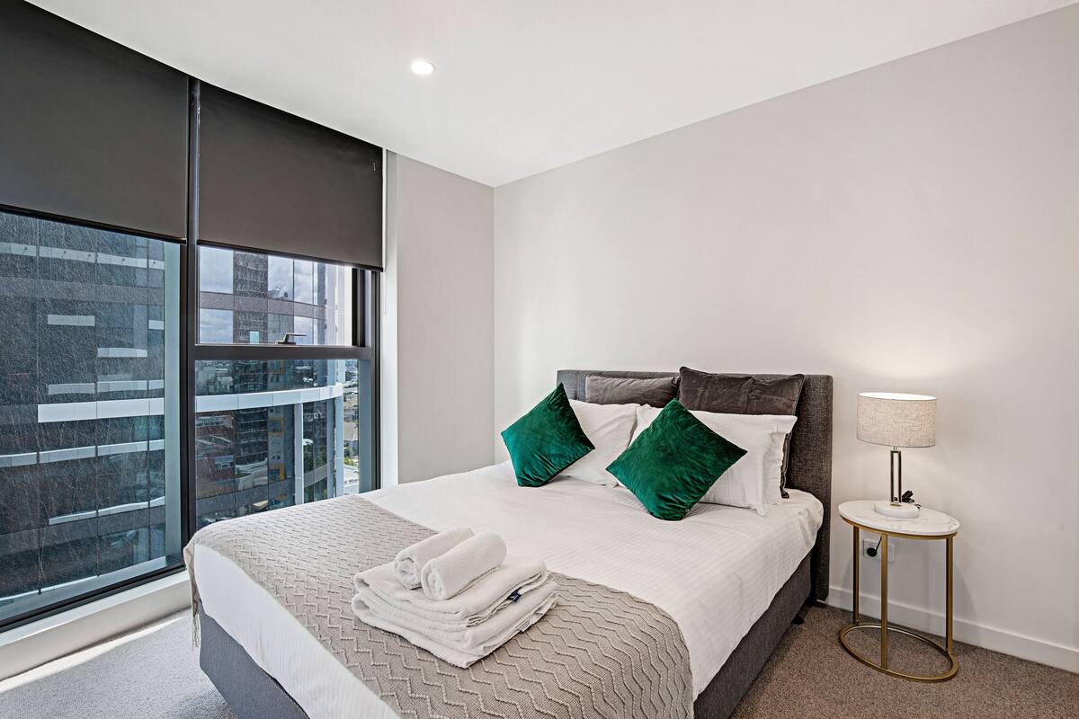Un hotel moderno con una cama grande, almohadas verdes, una lámpara blanca y vistas de la ciudad a través de ventanales de piso a techo.