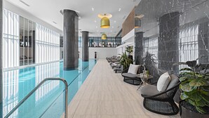 A modern indoor pool area with a glass barrier, lounge chairs, and potted plants. - Lumina Suites (West Melbourne)