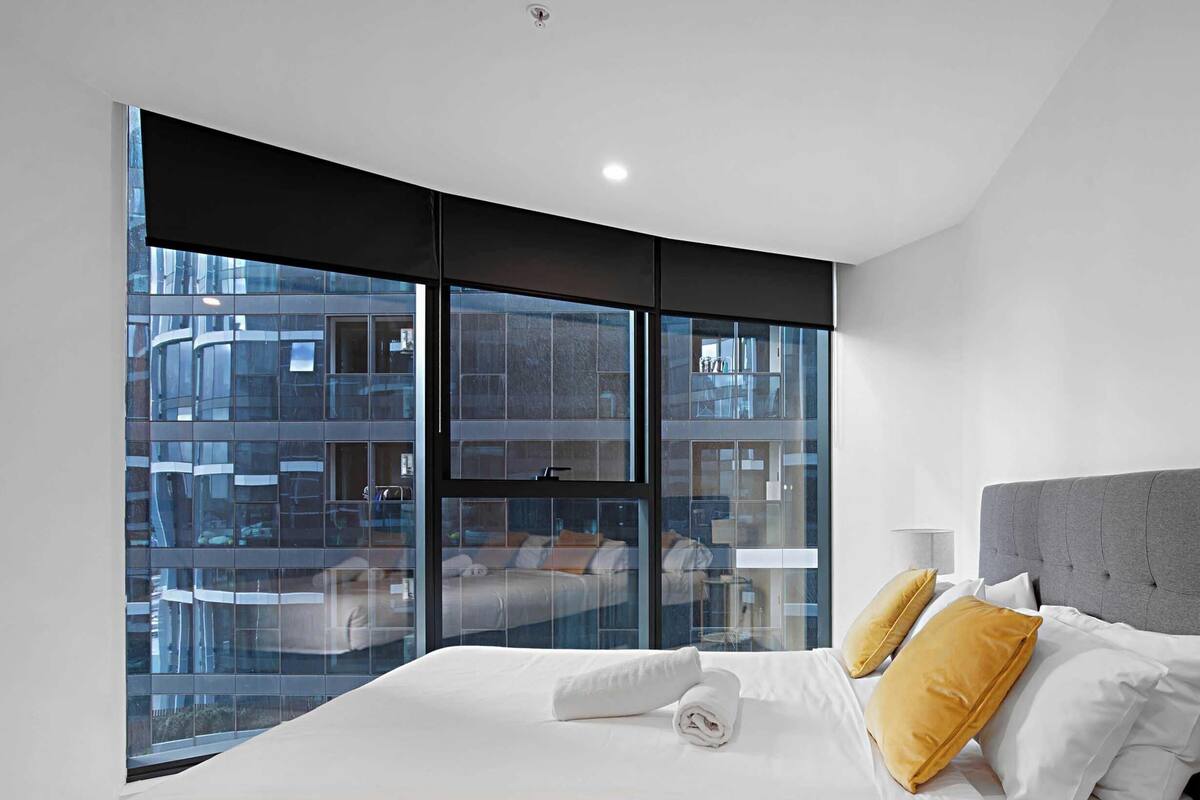 Una habitación de hotel moderna con un ventanal curvo, una cama grande con ropa blanca y un cabecero tapizado en gris.