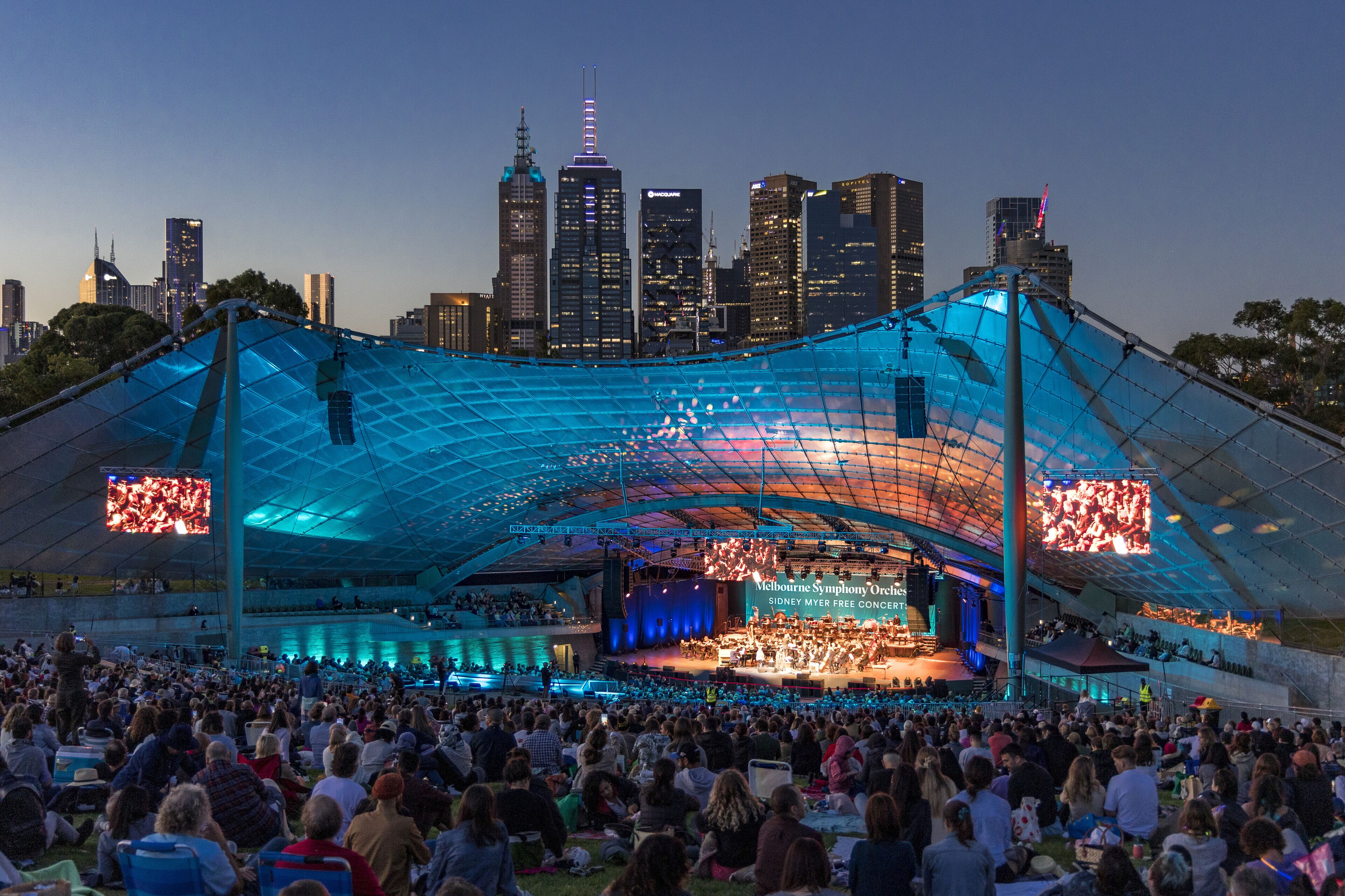 A large outdoor amphitheater with a stage and audience seating, set against a city skyline.