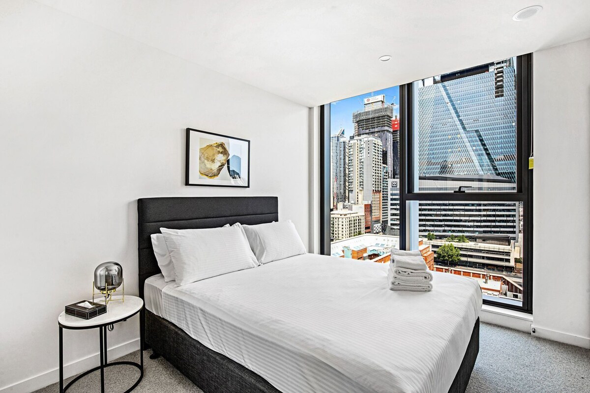 A hotel room with a large window, a bed with white bedding, a small bedside table with a lamp, and a framed picture on the wall.