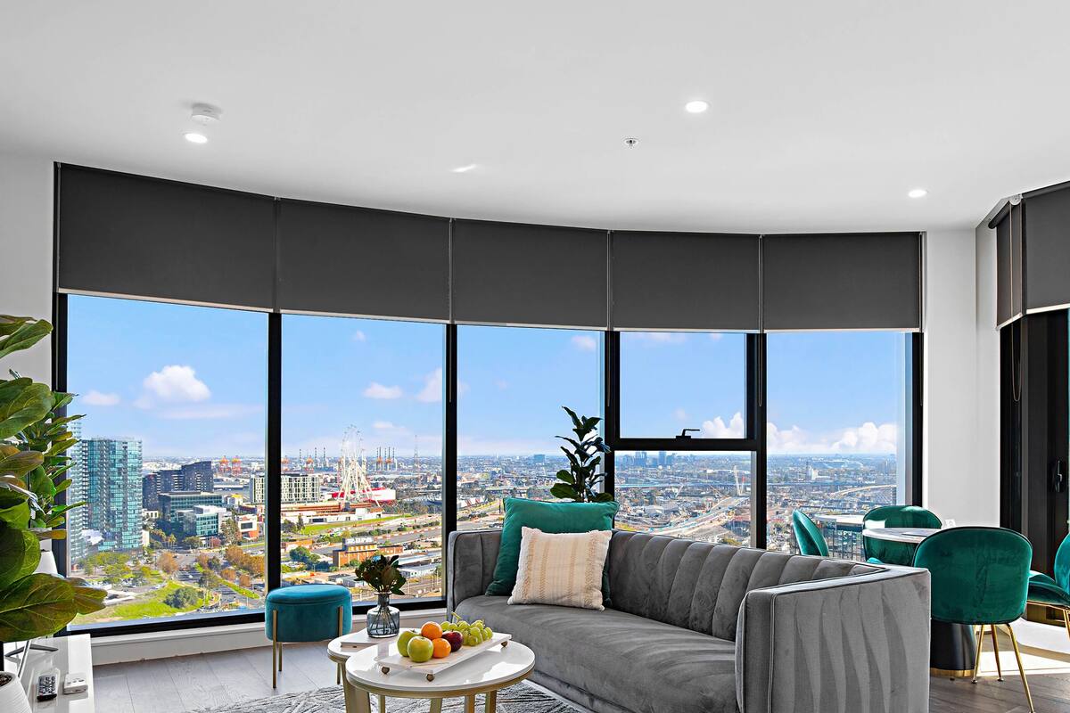 Una sala de estar moderna con un ventanal que ofrece vistas a la ciudad, un sofá gris, sillas color turquesa y una mesa de centro con fruta.