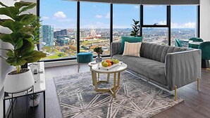 A modern living room with a large city view, a grey sofa, a round coffee table with fruit, and a potted plant. - Lumina Suites (West Melbourne)