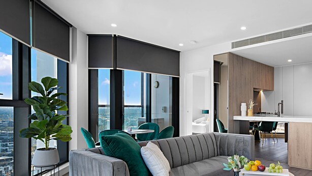 A modern living room with a grey sofa, teal chairs, a small round table, and a large window offering a city view. - Lumina Suites (West Melbourne)