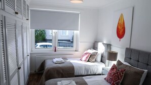 Een slaapkamer met twee bedden, een radiator, een raam met zonwering, een lamp en een aan de muur bevestigd kunstwerk.