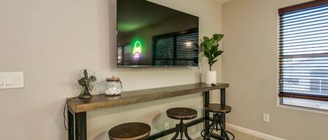 A modern living room with a flat-screen TV mounted on the wall, a wooden console table with three stools, a potted plant, and a decorative fan.