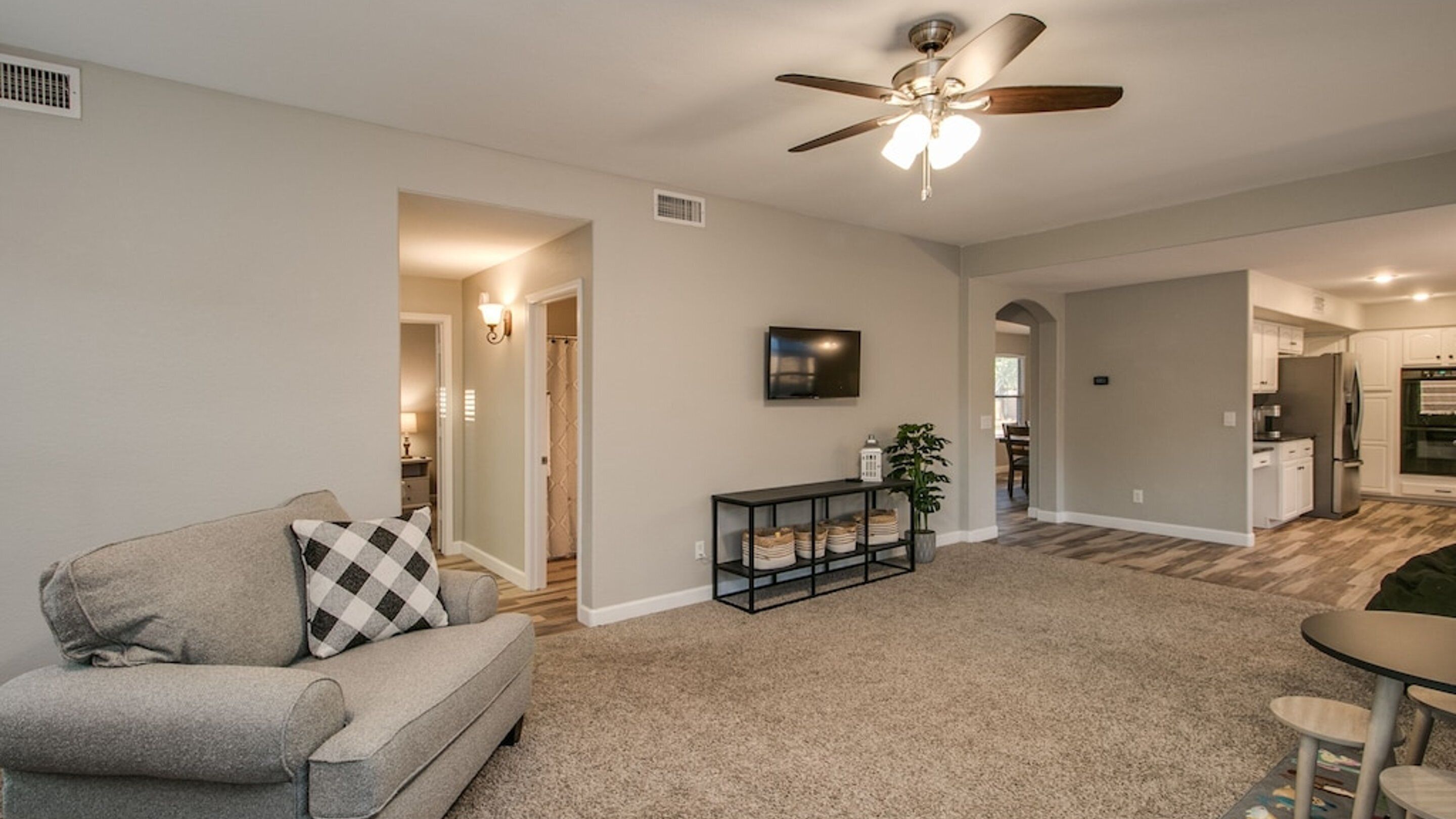 A living room with a grey sofa, a ceiling fan, a TV, and a kitchen in the background.