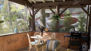 A covered outdoor seating area with wicker furniture, a grill, and a view of the surrounding trees and vehicles. - Boho shabby chic shack In beautiful resort (Newland)