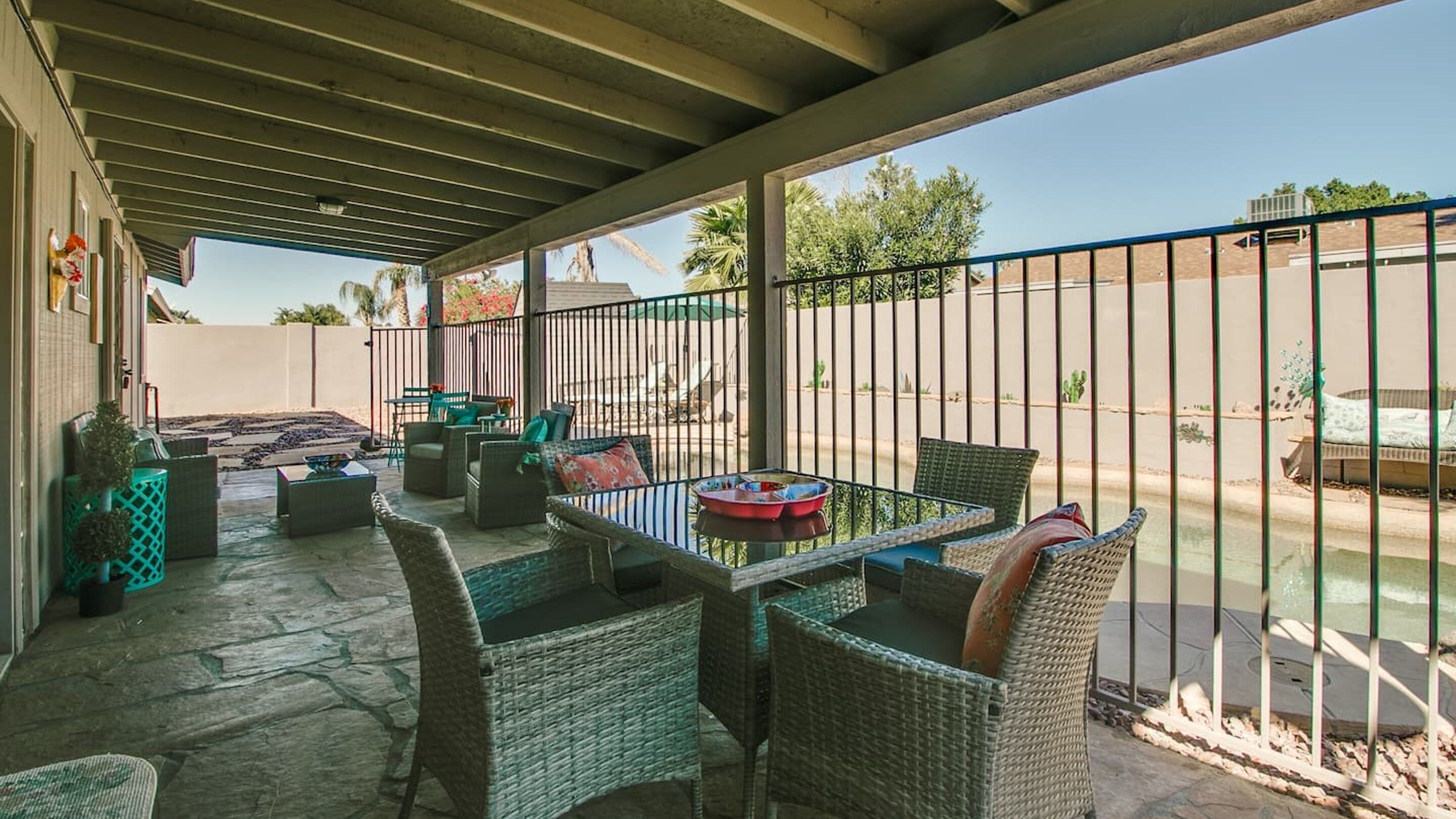 A covered patio with wicker furniture, a table, and chairs, overlooking a fenced yard with a pool.