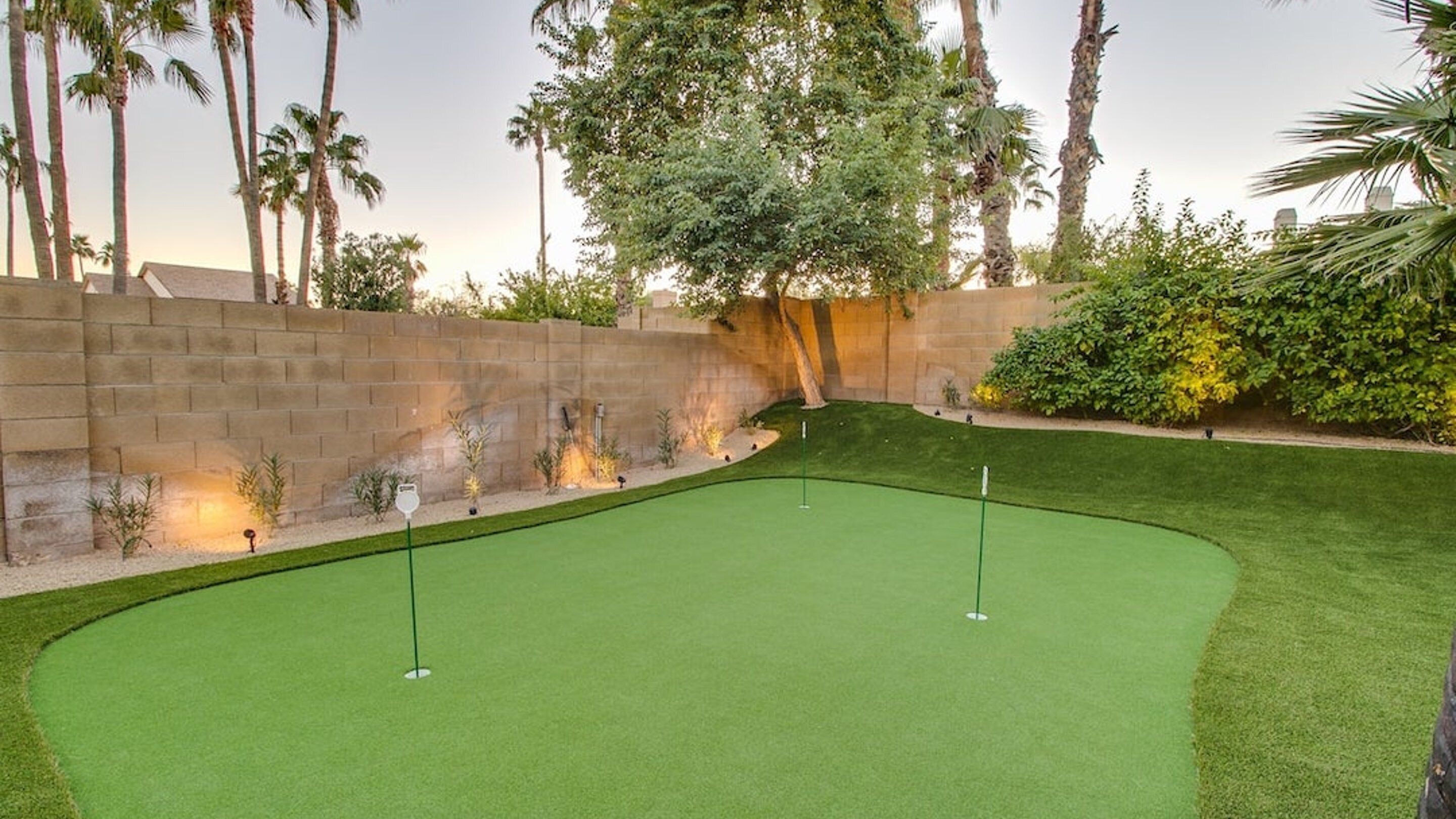 A well-maintained golf course with a putting green, surrounded by a stone wall and various plants.