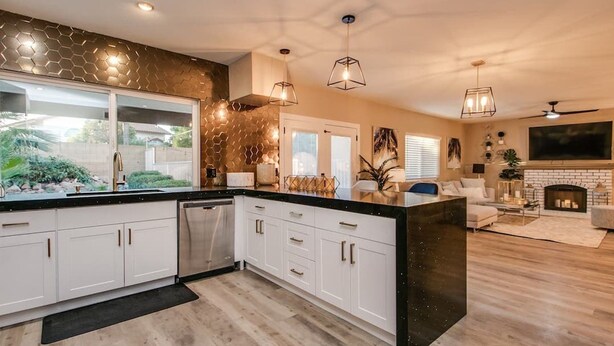 A modern kitchen with white cabinets, a black countertop, and a built-in dishwasher. There is a dining area with a white sofa and a fireplace. - Amazing Remodeled Scottsdale 5 Bdrm W/pool! (Scottsdale)